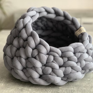 May include: A round, open-top basket constructed from thick, chunky grey fabric tubing woven in a braided pattern. The basket features a small, light-colored fabric tag on the interior rim with the text Cats&Dogs printed in black ink.