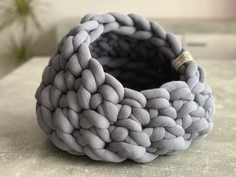 May include: A round, open-top basket constructed from thick, chunky grey fabric tubing woven in a braided pattern. The basket features a small, light-colored fabric tag on the interior rim with the text Cats&Dogs printed in black ink.