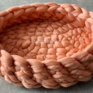 May include: A peach-colored, hand-knitted pet bed. The bed is round with a braided edge and a textured interior. The bed is made of thick, soft yarn, providing a cozy space for a small dog or cat. The website "catsanddogs.de" is visible on the side.