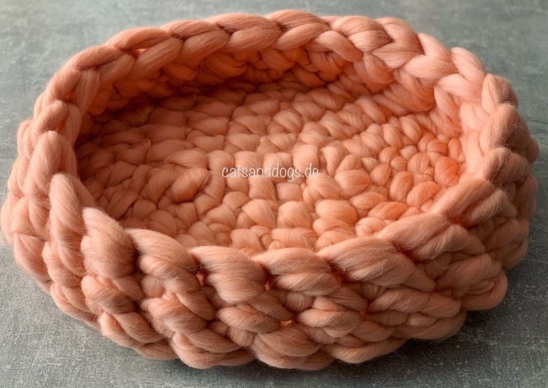 May include: A peach-colored, hand-knitted pet bed. The bed is round with a braided edge and a textured interior. The bed is made of thick, soft yarn, providing a cozy space for a small dog or cat. The website "catsanddogs.de" is visible on the side.