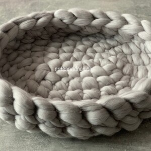 May include: A round, gray pet bed made of thick, braided wool. The bed has a low profile with a woven texture and a soft, inviting appearance. The text "catsanddogs.de" is visible on the side of the bed.