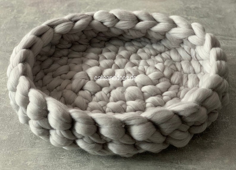 May include: A round, gray pet bed made of thick, braided wool. The bed has a low profile with a woven texture and a soft, inviting appearance. The text "catsanddogs.de" is visible on the side of the bed.