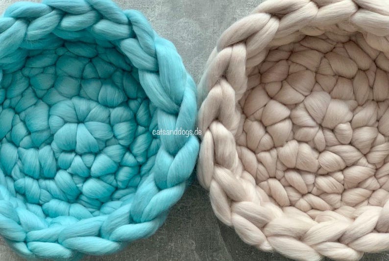 May include: Two round pet beds made of thick, chunky knit wool. One is a light teal color, and the other is a neutral beige. The beds have a braided edge and a textured interior, perfect for cats and dogs.