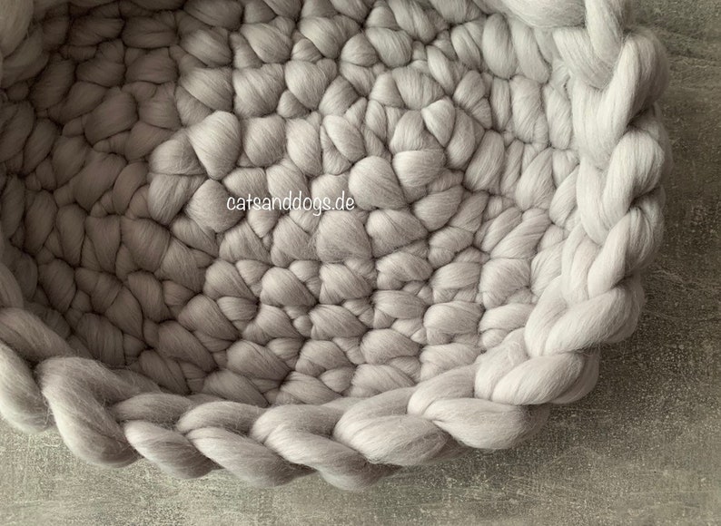 May include: A light gray, round pet bed made of thick, chunky wool. The bed has a braided edge and a textured interior. The website "catsanddogs.de" is visible in the image.