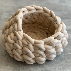 May include: A round, beige, chunky knit basket made from thick, braided fabric cord. The basket features a deep, open interior and a small, rectangular fabric tag on the rim with the text Cats&Dogs and a heart symbol printed in black ink.