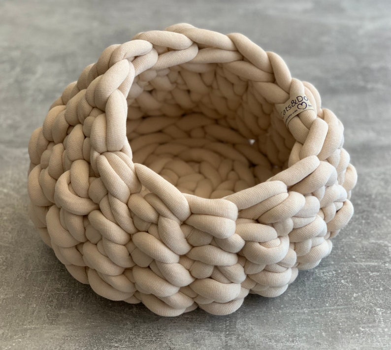 May include: A round, beige, chunky knit basket made from thick, braided fabric cord. The basket features a deep, open interior and a small, rectangular fabric tag on the rim with the text Cats&Dogs and a heart symbol printed in black ink.