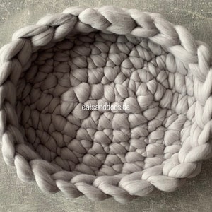 May include: A round, gray pet bed made of thick, woven wool. The bed has a braided edge and a textured interior. The website "catsanddogs.de" is visible on the lower right. The bed is designed for pets.
