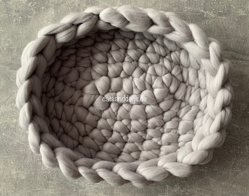 May include: A round, gray pet bed made of thick, woven wool. The bed has a braided edge and a textured interior. The website "catsanddogs.de" is visible on the lower right. The bed is designed for pets.