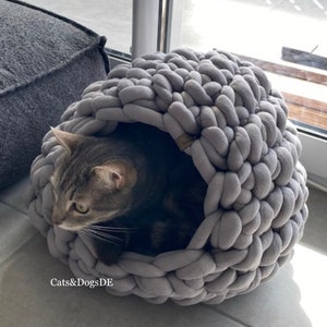 May include: A grey, chunky knit cat bed with a rounded, cave-like structure. A grey tabby cat is resting inside the soft, thick yarn enclosure. The text "Cats&DogsDE" is printed in white on the lower left side of the image.