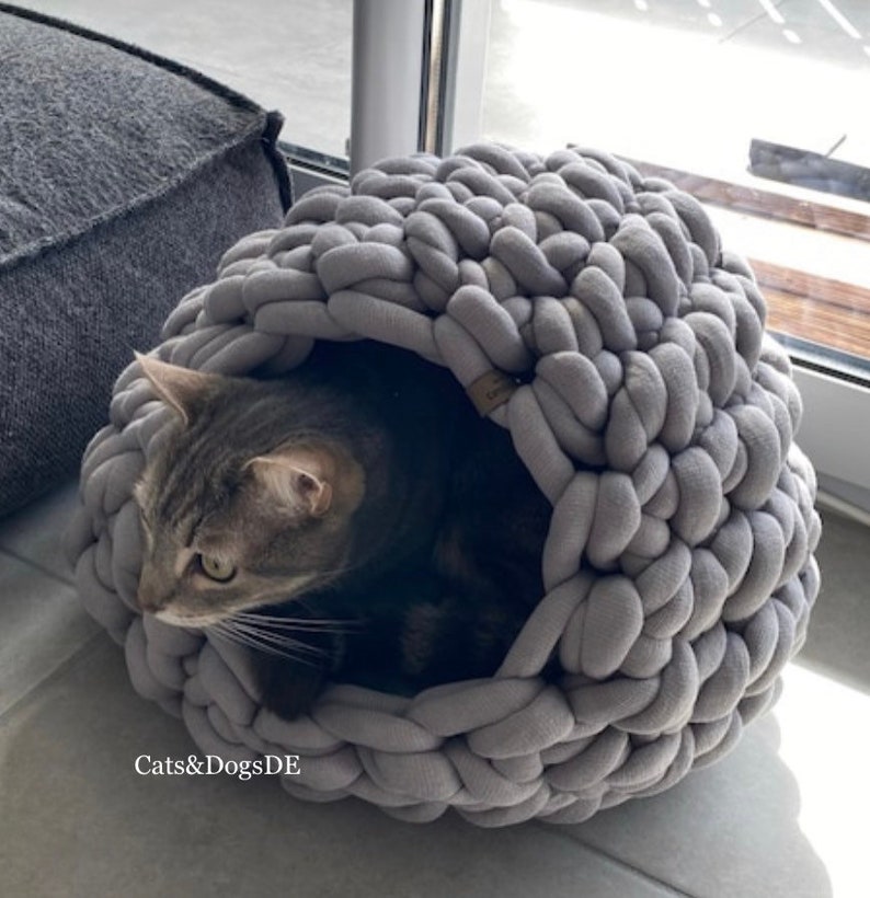 May include: A grey, chunky knit cat bed with a rounded, cave-like structure. A grey tabby cat is resting inside the soft, thick yarn enclosure. The text "Cats&DogsDE" is printed in white on the lower left side of the image.