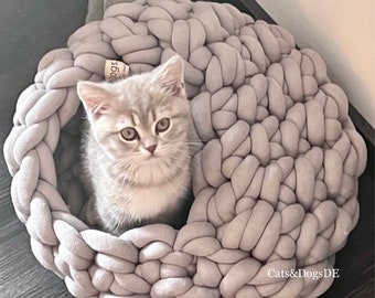 May include: A gray knitted pet bed with a cat sitting inside. The bed is made of thick, chunky yarn and has a round shape. The cat is white and gray with green eyes. The bed has the text "Cats&DogsDE" on the bottom.