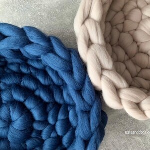 May include: Close-up of two chunky knit pet beds. One is a deep blue, the other is a light beige. The beds are circular with a braided edge. The texture of the wool is visible. The beds are on a light gray surface.