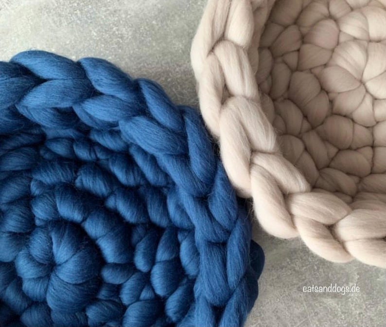 May include: Close-up of two chunky knit pet beds. One is a deep blue, the other is a light beige. The beds are circular with a braided edge. The texture of the wool is visible. The beds are on a light gray surface.