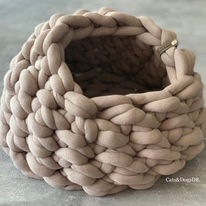 May include: A round, beige, chunky knit basket crafted from thick fabric cord. The basket features a wide, open top and a small, rectangular leather-like tag attached to the rim with a metal rivet, embossed with the text Cats&DogsDE.