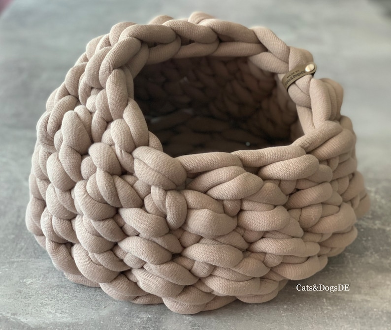 May include: A round, beige, chunky knit basket crafted from thick fabric cord. The basket features a wide, open top and a small, rectangular leather-like tag attached to the rim with a metal rivet, embossed with the text Cats&DogsDE.