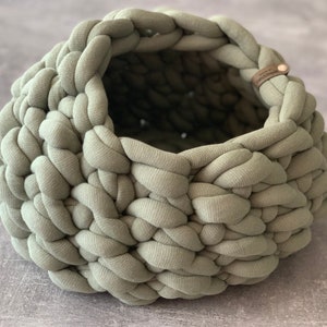 May include: A sage green round basket crafted from thick, chunky knit fabric. The basket features a wide, open top and a textured, braided appearance. A small, rectangular brown leather tag with the text "Crafts & Design" is attached to the rim.