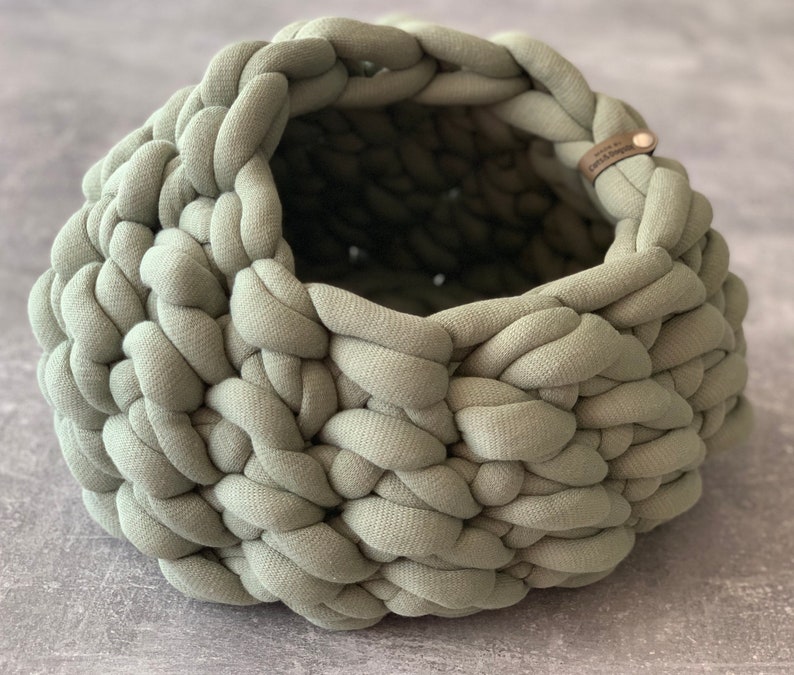 May include: A sage green round basket crafted from thick, chunky knit fabric. The basket features a wide, open top and a textured, braided appearance. A small, rectangular brown leather tag with the text "Crafts & Design" is attached to the rim.