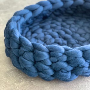 May include: A round, woven pet bed in a deep blue hue. The bed is made of thick, soft yarn, creating a textured, cozy appearance. The design features a braided edge and a spacious interior, suitable for small pets.