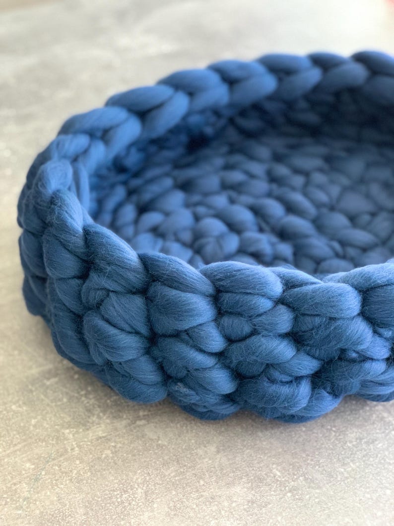 May include: A round, woven pet bed in a deep blue hue. The bed is made of thick, soft yarn, creating a textured, cozy appearance. The design features a braided edge and a spacious interior, suitable for small pets.