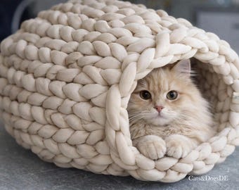 May include: A beige, woven cat bed shaped like a cozy cave. A fluffy, light-orange cat is nestled inside, peeking out with wide eyes. The bed is made of thick, braided yarn, creating a soft, inviting space for a pet.