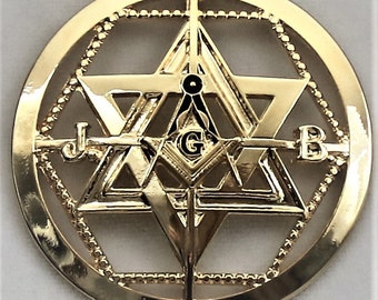 Könnte beinhalten: Goldfarbener Anhänger mit einem sechszackigen Stern, in dessen Mitte sich ein Zirkel und ein Winkelmaß befinden. Der Stern ist von einem Kreis mit einer hebräischen Inschrift und einem Perlenrand umgeben.