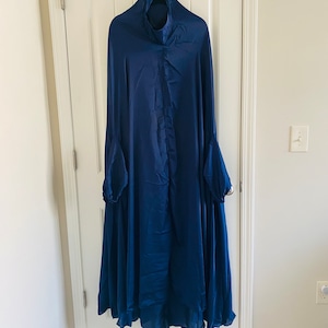 Puede incluir: Una chal de oración azul oscuro con capucha y mangas largas. La chal está hecha de una tela suave y fluida.