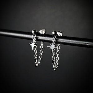 Puede incluir: Un par de pendientes de cadena plateados. Cada pendiente presenta un pequeño colgante de estrella colgando de una cadena. Los pendientes se muestran sobre un fondo oscuro, resaltando su brillo metálico. Los pendientes están diseñados para usarse a través de la oreja.
