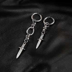 May include: A pair of silver hoop earrings with a dangling dagger charm. The earrings are on a black leather background.