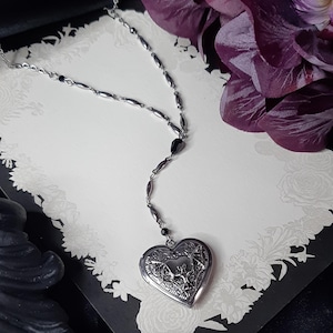 May include: A silver necklace with a heart-shaped locket pendant. The locket has an ornate design and hangs from a delicate chain with black bead accents. The necklace rests on a decorative card with floral patterns, next to a purple flower.