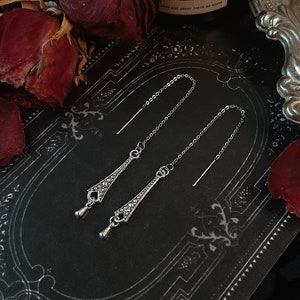 May include: A pair of silver dangle earrings with a delicate chain and a detailed design. The earrings are on a black background with a decorative border.
