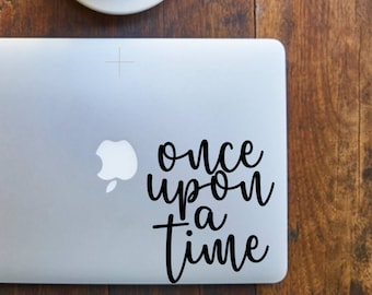 Może przedstawiać: Srebrny laptop z czarną naklejką z napisem "once upon a time" w prawym górnym rogu. Biały kubek do kawy z czarną kawą stoi na drewnianym stole.