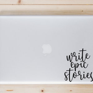 May include: A silver laptop with a black vinyl decal applied to the right side. The decal features the text "write epic stories" in a black, cursive, handwritten-style font. The laptop has a smooth, metallic finish and a centered apple logo.