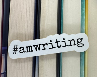 Op de afbeelding: Een witte sticker met zwarte tekst die "#amwriting" zegt, is op een stapel boeken geplakt.