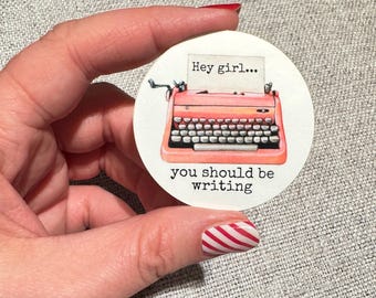 Puede incluir: Una pegatina redonda con una ilustración de una máquina de escribir rosa y el texto "Hey girl... you should be writing".