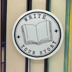 May include: White round sticker with a black border and the text "WRITE YOUR STORY" in a circular design. A black and white illustration of an open book is in the center of the sticker.