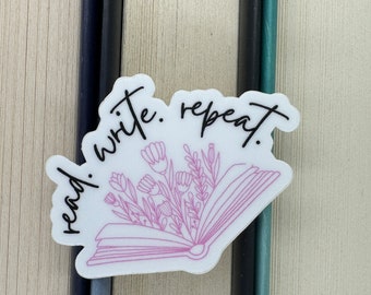 Op de afbeelding: Witte sticker met de tekst "read. write. repeat." in zwarte schrijfletters. Achter de tekst bevindt zich een roze omtrek van een open boek met bloemen erin.