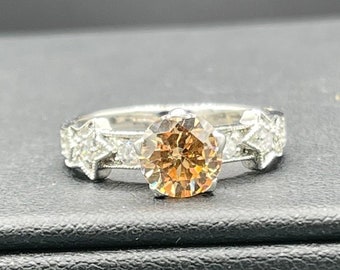 May include: A silver ring with a large, round, champagne-colored diamond in the center. Smaller diamonds are set on either side of the center stone.