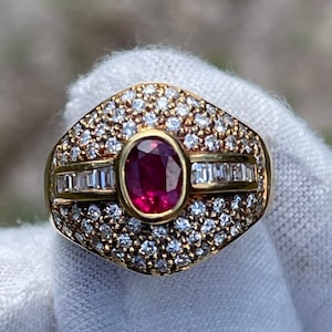 May include: A gold ring with a large oval ruby surrounded by smaller diamonds. The ring is set in a pave setting and has a polished finish.