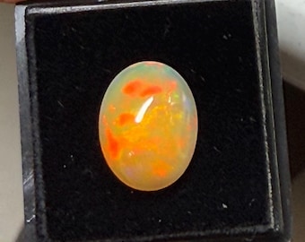 Pode incluir: Uma pequena gema de opala oval com cores laranja, amarela e branca em uma caixa de veludo preto. A caixa está etiquetada com "WHEE - 220".