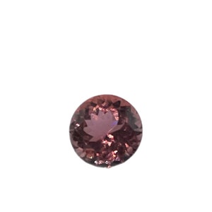 May include: A round, faceted gemstone in a deep rose color. The gem is set against a stark white background, with the word "BAZAART" in the bottom right corner.