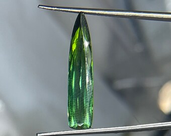 May include: A green gemstone with a pointed tip, held by tweezers. The gemstone has a faceted cut and a dark green colour.