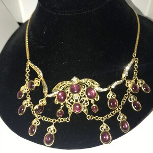 May include: A gold necklace with a delicate filigree design and a cluster of purple stones. The necklace has a dangling design with multiple strands of chain and purple stones.