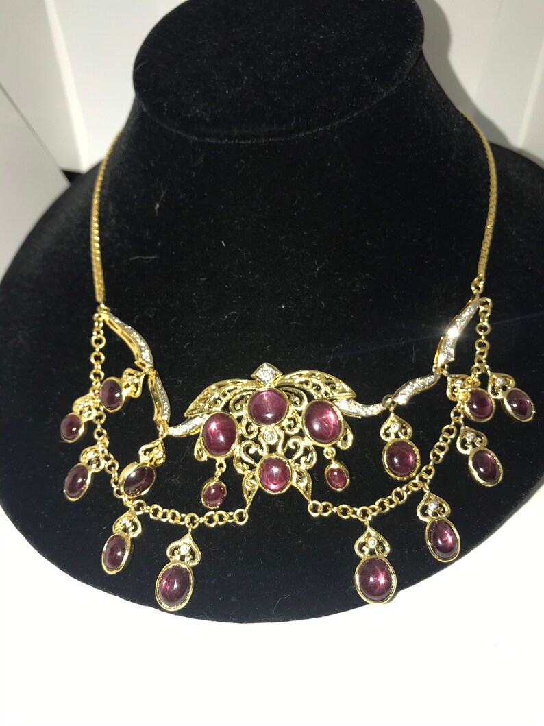 May include: A gold necklace with a delicate filigree design and a cluster of purple stones. The necklace has a dangling design with multiple strands of chain and purple stones.