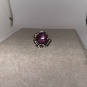 May include: A silver ring with a purple star ruby gemstone set in a decorative, starburst-shaped bezel.