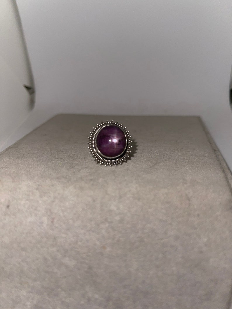 May include: A silver ring with a purple star ruby gemstone set in a decorative, starburst-shaped bezel.