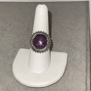 May include: A silver ring with a purple star-shaped gemstone set in a decorative bezel. The ring is on a white ring display stand.