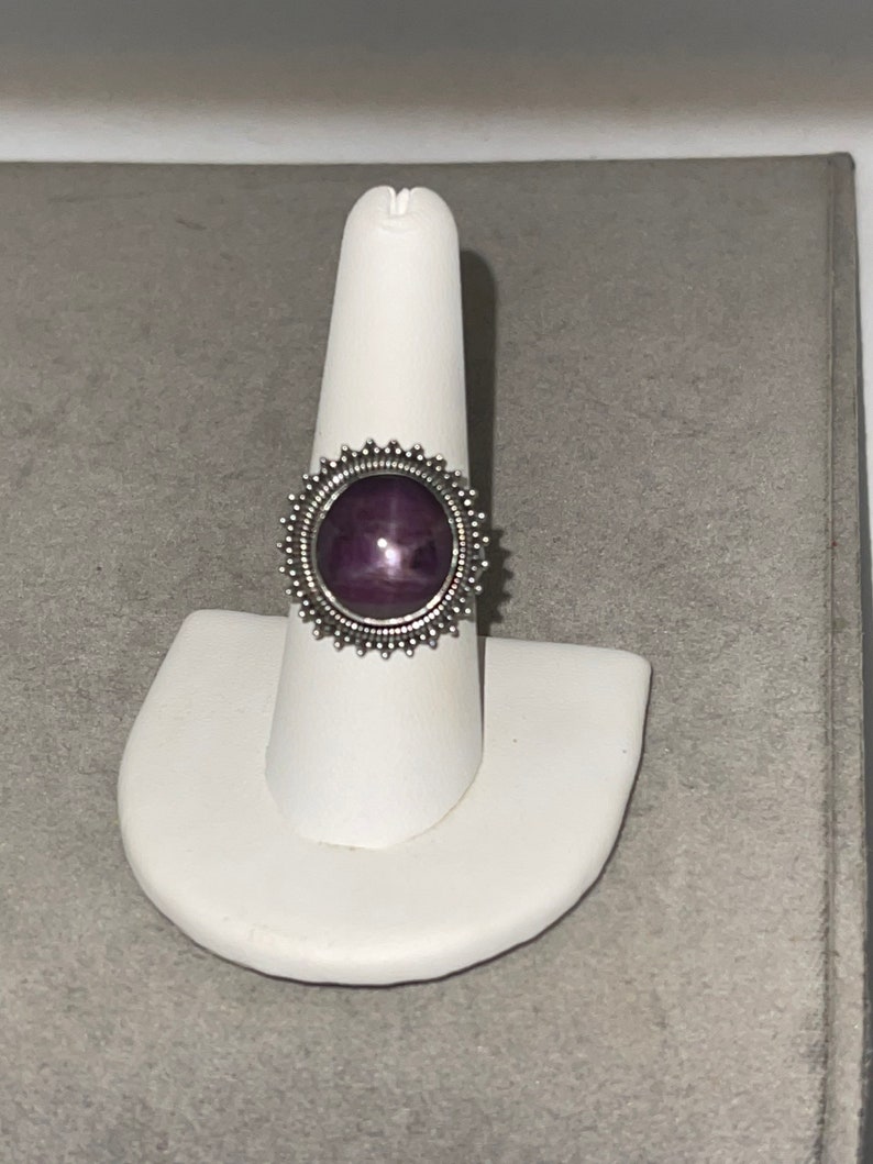May include: A silver ring with a purple star-shaped gemstone set in a decorative bezel. The ring is on a white ring display stand.