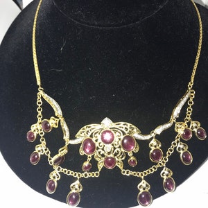 May include: A gold necklace with a delicate chain and a pendant featuring a floral design with purple gemstones. The pendant is surrounded by smaller purple gemstones that dangle from the chain.
