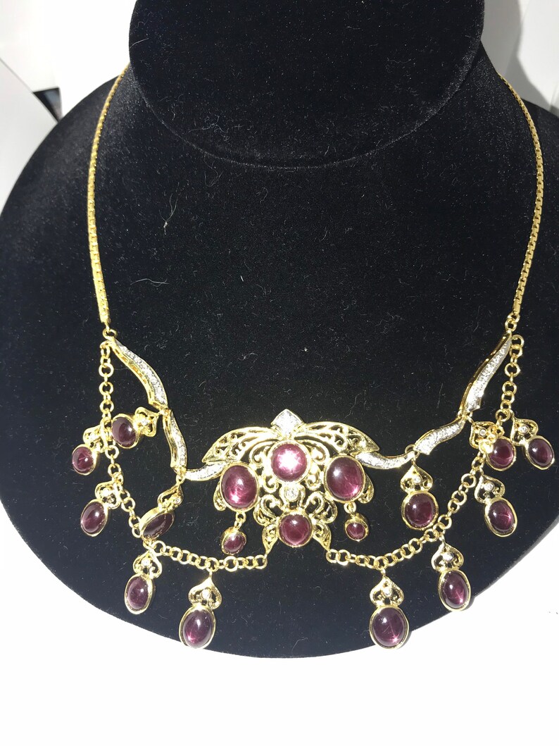 May include: A gold necklace with a delicate chain and a pendant featuring a floral design with purple gemstones. The pendant is surrounded by smaller purple gemstones that dangle from the chain.