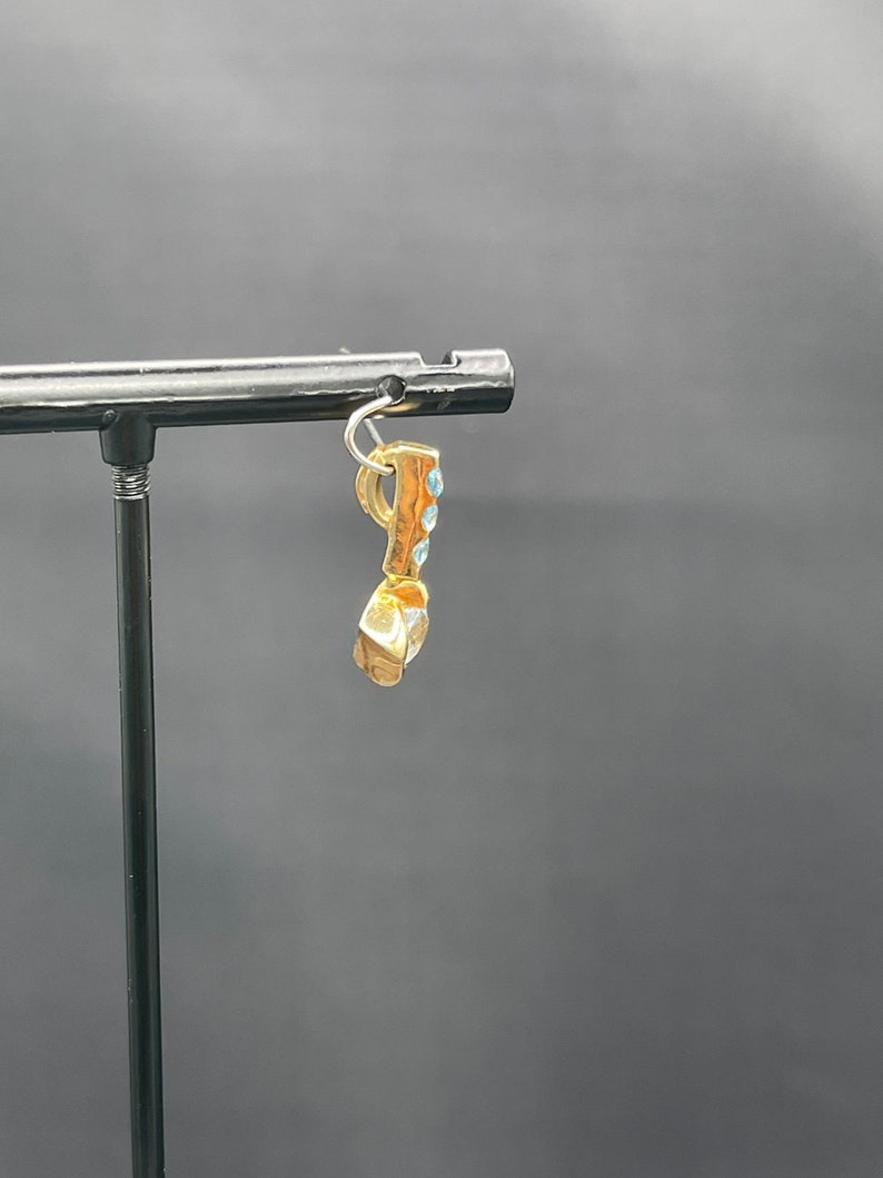 May include: Gold charm with blue stones, hanging from a black display stand.